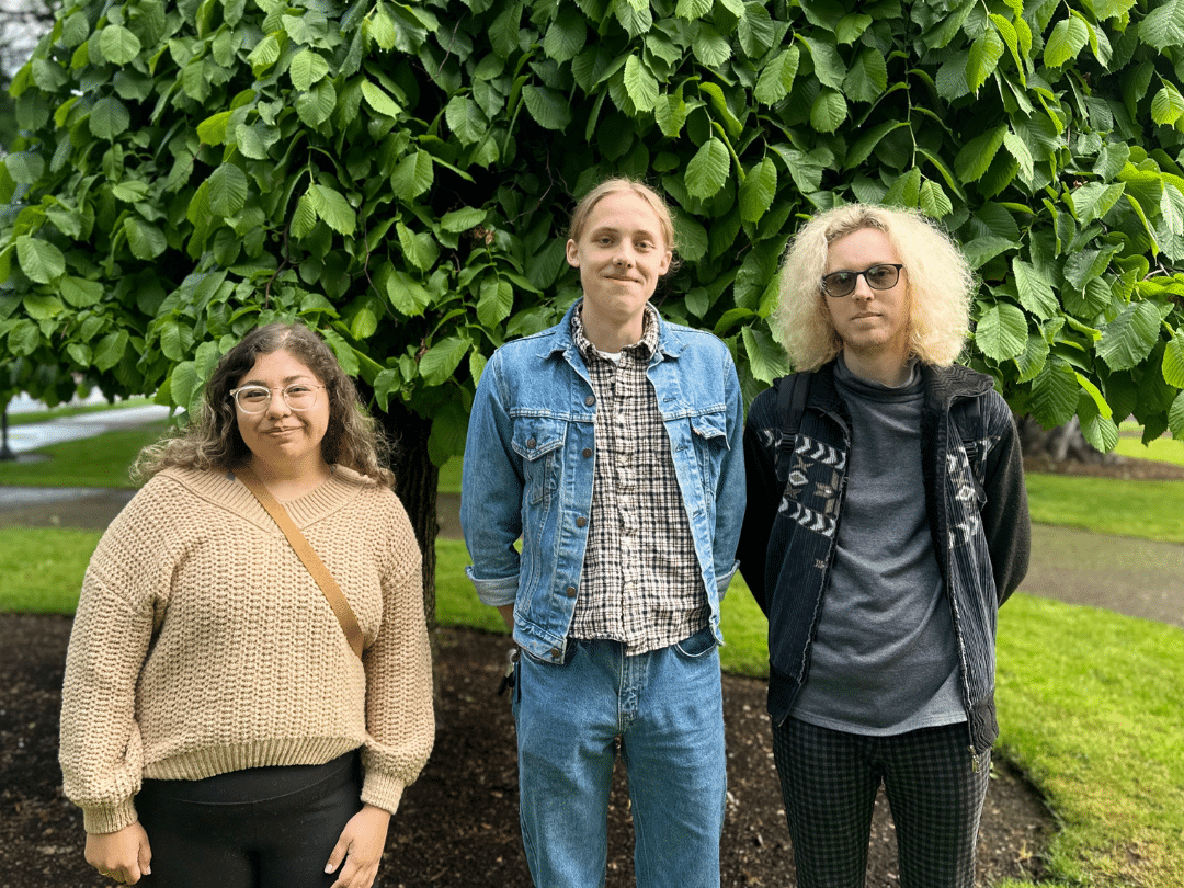 Three young adults stand side-by-side outdoors in front of a leafy green tree. From left to right: A person with shoulder-length dark hair and glasses wears a beige v-neck sweater. The person in the middle has light brown, shoulder-length hair and is wearing a blue denim jacket over a plaid shirt and jeans. The person on the right has curly, light blonde hair, sunglasses, a dark grey jacket with a geometric pattern, and plaid pants.