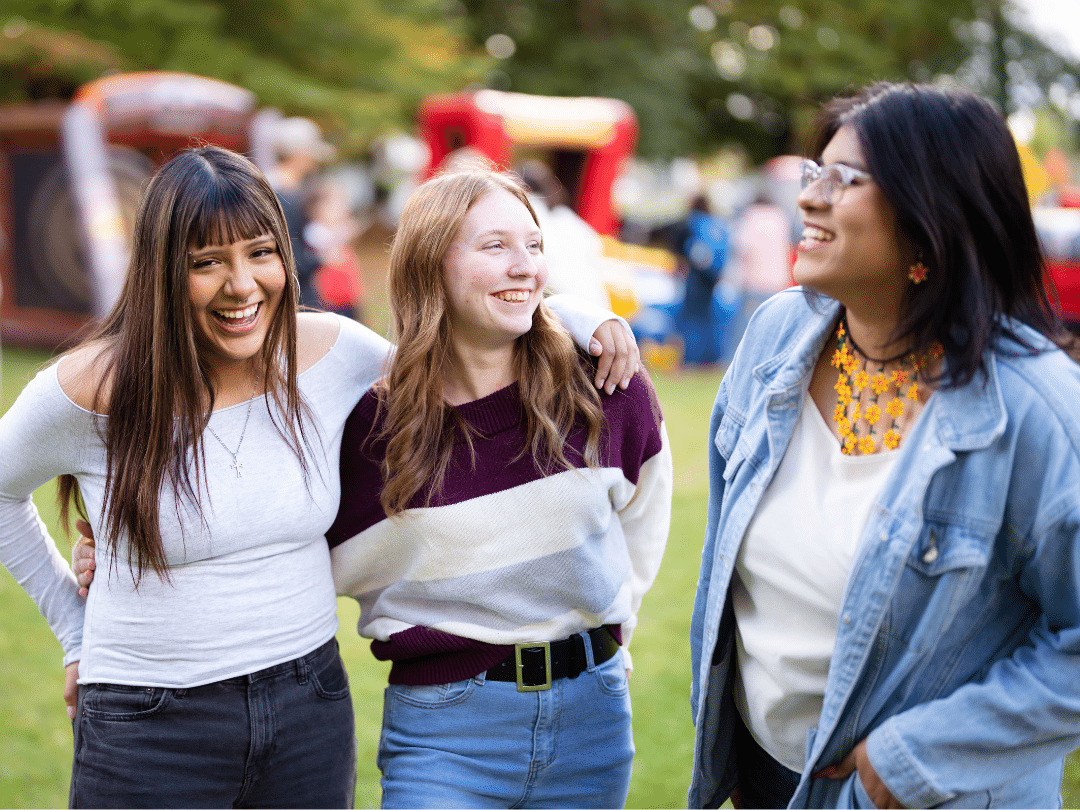 Three young women stand close together outdoors, smiling and laughing. The woman on the left has dark hair and is wearing a light-colored, off-the-shoulder top and dark jeans. The woman in the middle has auburn hair and is wearing a striped sweater with shades of purple, white, and tan, along with blue jeans. She has her arm around the woman on the left. The woman on the right has dark hair and is wearing a light-colored top under a denim jacket, with a bright yellow necklace. In the blurred background, there appears to be a park or outdoor event with inflatable structures.