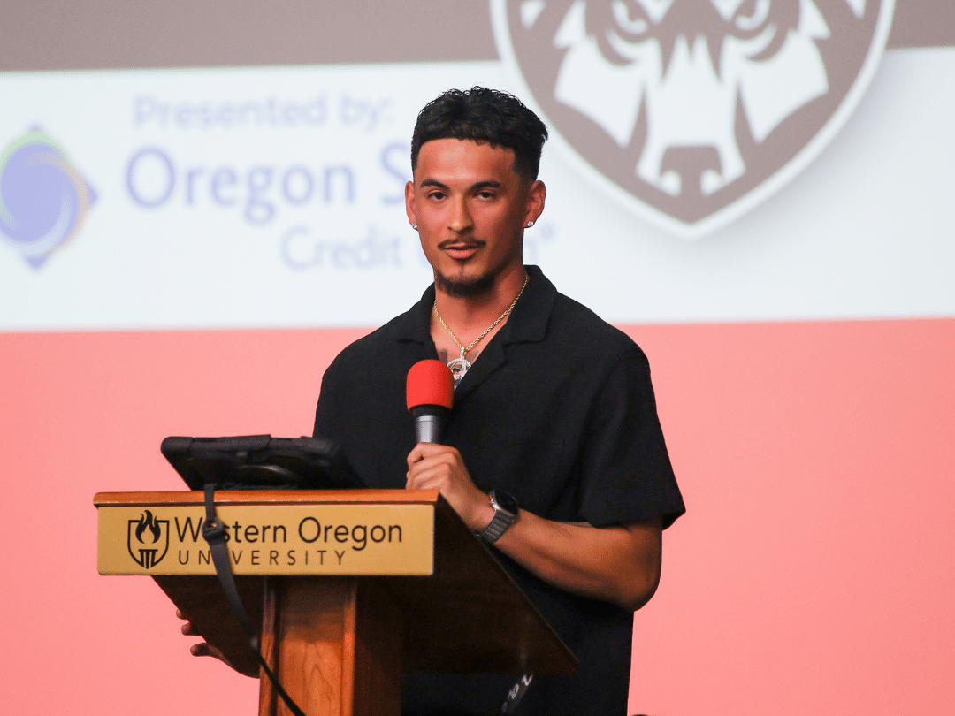 A man with short dark hair and a beard, wearing a black collared shirt and a necklace, stands at a wooden podium with 'Western Oregon University' inscribed on it. He holds a microphone in his right hand and looks forward. Behind him is a screen displaying 'Presented by: Oregon S Credit' and a stylized animal head logo.